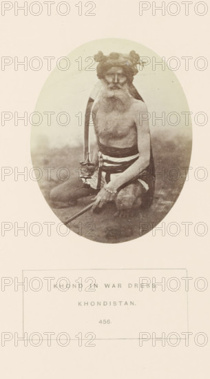 Portrait of an unknown man in his warrior clothes from the Khond tribe in Odisha, India, photograph, page, number: '456.', inscription: 'KHOND IN WAR DRESS., KHONDISTAN.', photographer: Charles Philip Yorke Triscott (possibly), Odisha, c. 1865 - in or before 1875, East India, paper, albumen print, height 98 mm x width 77 mm, clothes, costume, human races, peoples, nationalities, A sepia-toned oval photograph shows an older bearded man seated on the ground with bare torso and a cloth wrapped around his lower body, a long curved object resting beside him and a straight staff held in his hands, a cloth or headwrap with decorative elements on his head, a bracelet on one wrist, and a plain light background surrounding the figure.