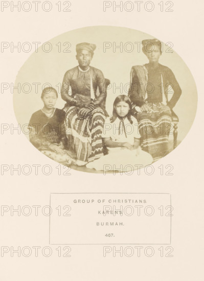 Group portrait of four unknown people from the Karen folk of Myanmar, photograph, page, inscription: 'GROUP OF CHRISTIANS., KARENS., BURMAH.', number: '467.', Myanmar, c. 1865 - in or before 1875, photographer, paper, albumen print, height 95 mm x width 120 mm, human races, peoples, nationalities, A faded oval photograph shows four seated people framed by a pale background; two adults sit on chairs in the back wearing dark long-sleeved tops and patterned lower garments with headwraps, hands resting in their laps, while two younger individuals sit in front on the floor wearing lighter and darker clothing, one in a light dress with a ribbon at the collar and the other in a darker outfit with a patterned skirt; all faces are visible with neutral expressions and dark hair, and the image has a soft, sepia-toned appearance.