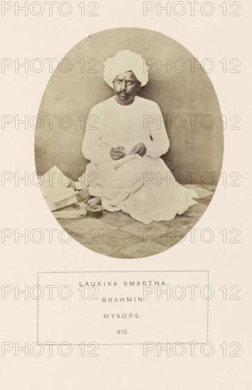 Portrait of an unknown priest from the Smarta Brahmin community of Mysore, Karnataka, India, photograph, page, number: '412.', inscription: 'LAUKIKA SMARTHA., BRAHMIN., MYSORE.', Mysore, c. 1865 - in or before 1875, photographer, South India, paper, albumen print, height 101 mm x width 87 mm, Brahma (supreme being, creator, etc., Hinduism, Buddhism, Jainism), human races, peoples, nationalities, An oval-framed sepia photograph of a man seated on a patterned floor, wearing a light-colored turban and matching long-sleeved garment with draped cloth around his legs; he holds a small object between his hands and gazes forward with a neutral expression; beside him on the floor are several folded papers or books and a round container, and the background is plain and softly shaded.