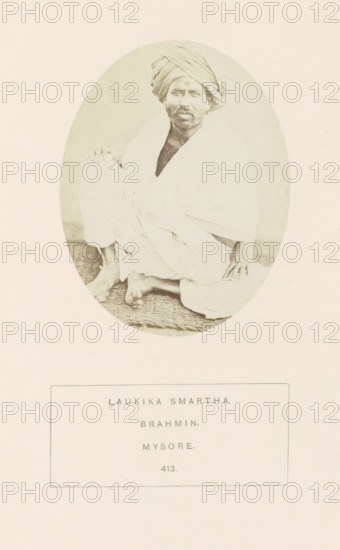 Portrait of an unknown priest from the Smarta Brahmin community of Mysore, Karnataka, India, photograph, page, number: '413.', inscription: 'LAUKIKA SMARTHA., BRAHMIN., MYSORE.', Mysore, c. 1865 - in or before 1875, photographer, South India, paper, albumen print, height 98 mm x width 76 mm, head-gear: turban, Brahma (supreme being, creator, etc., Hinduism, Buddhism, Jainism), human races, peoples, nationalities, An oval-framed sepia photograph shows a seated man wearing a wrapped turban and draped cloth garments, sitting cross-legged on a textured mat with one hand resting on his knee and the other on the mat; his face has a beard and he looks toward the camera, and the image is centered on a pale background with a rectangular bordered area below the oval.
