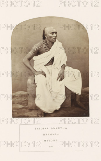 Portrait of an unknown priest from the Smarta Brahmin community of Mysore, Karnataka, India, photograph, page, inscription: 'VAIDIKA SMARTHA., BRAHMIN., MYSORE.', number: '414.', Mysore, c. 1865 - in or before 1875, photographer, South India, paper, albumen print, height 123 mm x width 95 mm, Brahma (supreme being, creator, etc., Hinduism, Buddhism, Jainism), human races, peoples, nationalities, A sepia-toned photograph of a seated man dressed in light-colored draped cloth, with one shoulder exposed and the fabric wrapped around his lower body and legs, sitting on a rectangular stone block with one foot on the floor and the other foot tucked to the side; he has a shaved head with a small patch of hair at the back, wears a beaded necklace and earrings, and his arms and torso show horizontal markings; his gaze is directed slightly to the side, and the background is plain and neutral.