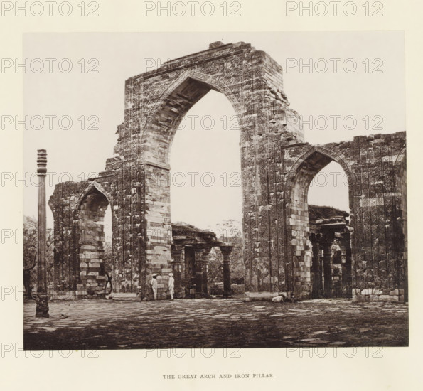 View of a gate and pillar at the Qutb complex in Delhi, The great arch and iron pillar, page, photomechanical print, photographer: Bourne & Shepherd, Delhi, c. 1860 - c. 1877, printmaker, paper, Woodburytype, height 146 mm x width 177 mm, arch, archivolt, architecture, column, pillar, A sepia-toned photograph of a large ornate stone archway and adjacent ruins, showing detailed carved patterns along the arch and supporting pillars, with visible masonry blocks and decorative motifs on vertical surfaces; a tall slender column stands to the left of the arch, and smaller arches and stone structures are visible through and beside the main arch; two figures in light clothing stand beneath the arch near its base; the ground in front is paved with irregular stone slabs and scattered stones, and trees and vegetation are visible through openings among the ruins.
