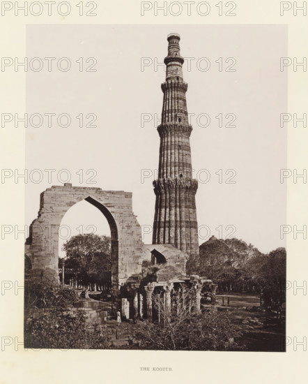 View of the Qutb Minar in Delhi, The Kootur, page, photomechanical print, photographer: Bourne & Shepherd, Delhi, c. 1860 - c. 1877, printmaker, paper, Woodburytype, height 182 mm x width 145 mm, tower, clock tower, monumental adornment, A tall, cylindrical stone tower with carved horizontal bands and decorative balconies rises behind a partially ruined stone archway, both set among trees and scattered stone ruins; the foreground shows smaller stone structures with pillars and vegetation, and a few small human figures near the base of the ruins.