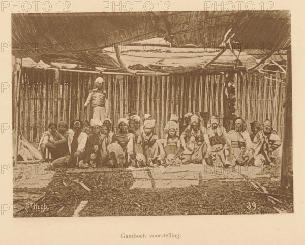 Portrait of unknown persons in costume, some with masks, Gamboeh scene, page, photomechanical print, maker: Christiaan Johan Neeb, c. 1892 - in or before 1897, paper, collotype, photoprint, height 118 mm x width 163 mm, mask, anonymous historical persons depicted in a group, in a group-portrait, stage costume, Indonesia, A sepia-toned photograph showing a group of people seated and kneeling on a floor in front of a wall made of vertical bamboo slats, with a thatched roof overhead supported by wooden poles and beams. The people wear a variety of clothing and head coverings, some holding round handheld fans and some wearing masks or face adornments; one person stands behind the seated group near the center. The ground in front of the group appears covered with woven mats and scattered natural debris, and the scene is framed by the rough wooden structure and roof elements above.