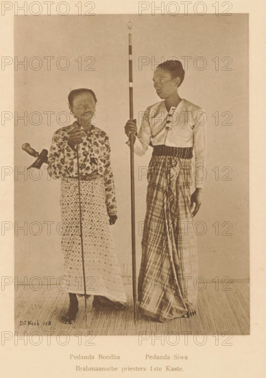 Brahmin priests 1st Caste, Portrait of Pedanda Buddha and Pedanda Siwa, page, photomechanical print, maker: Christiaan Johan Neeb, c. 1892 - in or before 1897, paper, collotype, photoprint, height 166 mm x width 123 mm, historical persons, adult man, Indonesia, A monochrome photograph of two standing people on a plain backdrop, both barefoot on a textured floor. The person on the left is shorter, wearing a patterned long skirt and a floral long-sleeved blouse, holding a thin vertical staff and a small object at their side, with short hair and a neutral facial expression. The person on the right is taller, wearing a light-colored long-sleeved top with decorative fastenings and a checked wrapped lower garment, holding a tall striped pole that reaches above their head, with hair pulled back and looking slightly toward the left. The overall image has a sepia tone and shows both figures in full length.