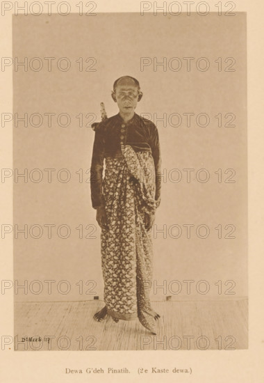 Portrait of Dewa G'deh Pinatih, Dewa G'deh Pinatih (2nd Caste dewa), page, photomechanical print, maker: Christiaan Johan Neeb, c. 1892 - in or before 1897, paper, collotype, photoprint, height 170 mm x width 119 mm, historical persons, adult man, Indonesia, A sepia-toned photograph of a standing person facing forward wearing a patterned wrap skirt and a long-sleeved dark top with decorative buttons, a folded patterned cloth draped across the torso, and a sheathed dagger tucked behind the shoulder; the person has a shaved or closely cropped head, visible ears, and a neutral facial expression, and the image shows a simple plain background and a textured floor beneath their feet.