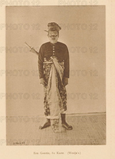 A Goestie, 3rd Caste. (Wesija's), Portrait of an unknown man, page, photomechanical print, maker: Christiaan Johan Neeb, c. 1892 - in or before 1897, Christiaan Johan Neeb, paper, collotype, photoprint, height 170 mm x width 119 mm, historical persons, adult man, Indonesia, A sepia-toned photograph of a barefoot man standing against a plain backdrop, wearing a patterned headwrap, a dark long-sleeved buttoned shirt, and a patterned cloth wrapped around his lower body with one end draped across his torso; a sheathed dagger with a visible handle is tucked at his side, and his hands hang at his sides while his feet rest on a textured floor.