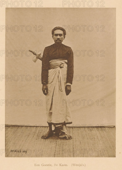 Portrait of an unknown man, A Goestie, 3rd Caste. (Wesija's), page, photomechanical print, maker: Christiaan Johan Neeb, c. 1892 - in or before 1897, Christiaan Johan Neeb, paper, collotype, photoprint, height 170 mm x width 119 mm, historical persons, adult man, Indonesia, A sepia-toned photograph of a person standing against a plain backdrop wearing a dark long-sleeved top and a light wrapped lower garment tied at the waist, with bare feet visible and one foot partially resting on a patterned cloth; the person has short, combed-back hair and a neutral facial expression, and a decorated handle of an object protrudes from the waist area on one side.
