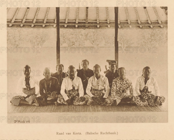 Group portrait of unknown men, the Council of Kerta, Council of Kerta (Balian Court.), page, photomechanical print, maker: Christiaan Johan Neeb, c. 1892 - in or before 1897, paper, collotype, photoprint, height 122 mm x width 167 mm, historical persons, adult man, Indonesia, A sepia-toned photograph showing a row of men seated on a woven mat under a roof with exposed beams and vertical posts, the men wearing patterned cloths wrapped around their lower bodies and a variety of shirts and jackets, some with patterned tops and some plain, several of the men holding or positioned near curved-handled objects, the background behind them appearing light and textured with faint silhouettes of trees, the floor in front of them textured and the scene framed by the roof structure above.