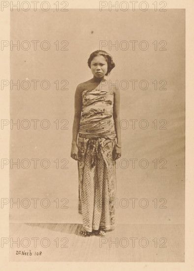 Bali woman from the 4th Caste. (Soendra's), Portrait of an unknown woman, page, photomechanical print, maker: Christiaan Johan Neeb, c. 1892 - in or before 1897, Christiaan Johan Neeb, paper, collotype, photoprint, height 117 mm x width 167 mm, historical persons, adult woman, Indonesia, A full-length, sepia-toned photograph of a young woman standing against a plain backdrop, wearing a patterned wrap around her chest and a long patterned skirt that reaches the floor, with her arms relaxed at her sides and her bare feet visible at the bottom, her hair styled back and a neutral expression on her face.
