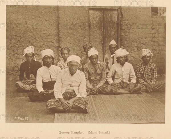 Guru Bangkol. (Mami Ismail), Group portrait of nine unknown men, page, photomechanical print, maker: Christiaan Johan Neeb, c. 1892 - in or before 1897, paper, collotype, photoprint, height 121 mm x width 166 mm, historical persons, adult man, anonymous historical persons depicted in a group, in a group-portrait, Indonesia, A sepia photograph showing a group of people seated on a woven mat in front of a textured wall and a wooden door, most wearing cloth headwraps and patterned lower garments, with several wearing light-colored shirts and some wearing darker patterned tops, all sitting closely together with hands resting in laps or on knees, faces turned toward the camera and showing neutral expressions, the scene framed by the rough wall surface on either side and the mat extending in front of them.