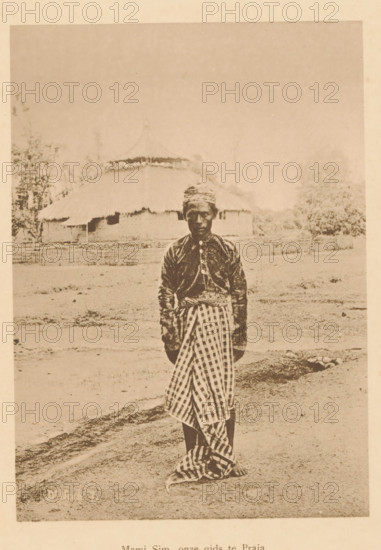 Mami Sim, our guide to Praja, Portrait of Mami Sim, page, photomechanical print, maker: Christiaan Johan Neeb, c. 1892 - in or before 1897, paper, collotype, photoprint, height 169 mm x width 120 mm, historical persons, adult man, Indonesia, A sepia-toned photograph of a person standing outdoors on bare ground, wearing a patterned headwrap, a dark long-sleeved top with buttons down the front, and a checkered cloth wrapped around the lower body, with bare feet visible; behind the person is a low fence and a thatched-roof building with a rounded roofline, surrounded by trees and open space.