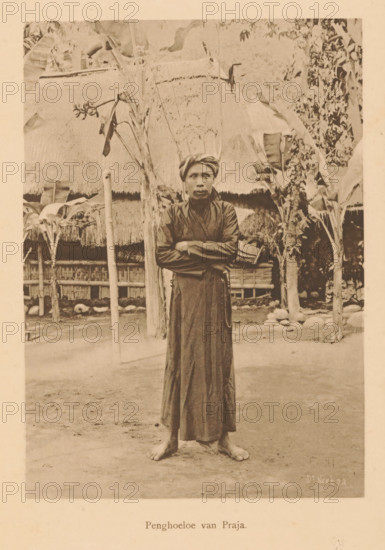 Portrait of an unknown man who is an Islamic teacher, Pengoeloe van Praja, page, photomechanical print, maker: Christiaan Johan Neeb, c. 1892 - in or before 1897, paper, collotype, photoprint, height 161 mm x width 118 mm, historical persons, adult man, Indonesia, A sepia-toned photograph of a barefoot person standing on bare ground with arms crossed, wearing a long dark robe and a wrapped headscarf, positioned centrally in front of a thatched structure and flanked by tall plants with broad leaves; behind them are wooden posts, woven fencing, and clustered stones, with foliage and parts of the thatched roof visible across the upper area of the image.
