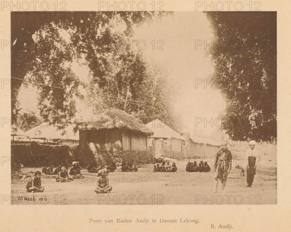 Poeri van Raden Andji te Dassan Lekong, Unknown persons, sitting and standing, at various houses, page, photomechanical print, maker: Christiaan Johan Neeb, c. 1892 - in or before 1897, paper, collotype, photoprint, height 122 mm x width 163 mm, historical persons, other dwelling types, Indonesia, A faded sepia photograph showing a dirt open area in front of simple thatched and walled structures, with large trees rising behind them and a pale sky above; people are seated on the ground in small groups across the foreground and midground, a few individuals sit alone with legs crossed, and two men stand near the right edge beside a dog, one wearing loose garments and the other in lighter clothing; stone or earthen walls and clustered huts form a low row behind the seated figures.