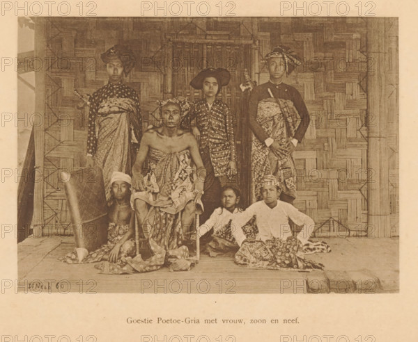 Goestie Poetoe-Gria with wife, son and nephew, Group portrait of a Goestie Poetoe-Gria with six unknown persons, page, photomechanical print, maker: Christiaan Johan Neeb, c. 1892 - in or before 1897, paper, collotype, photoprint, height 166 mm x width 120 mm, historical persons, adult woman, adult man, Indonesia, A sepia photograph shows a group of people arranged in front of a woven bamboo wall and a doorway. Some individuals are seated on a low platform while others stand behind them; several wear patterned cloth wrapped around their bodies and head coverings of different shapes and styles. One seated person in the center sits on a chair with cloth draped over the lap and shoulders, another seated figure beside them wears a lighter-colored top and patterned lower garment, and a few younger figures sit or crouch on the floor with cloths around their waists. The standing figures wear long patterned wraps and head coverings, and one holds a long object across the torso. The floor shows...