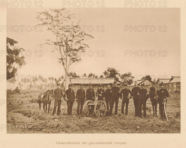 Group portrait of unknown officers near a cannon, Non-commissioned officers of the combined troops, page, photomechanical print, maker: Christiaan Johan Neeb, c. 1892 - in or before 1897, paper, collotype, photoprint, height 122 mm x width 169 mm, firearms: cannon, anonymous historical persons depicted in a group, in a group-portrait, ranks (in the army, navy, air force), A sepia-toned photograph shows a group of uniformed men standing in a line outdoors on grassy ground, some holding objects at their sides and two positioned beside horses, with a small wheeled cart in front of them; behind them are low wooden buildings and scattered trees, including a tall slender tree rising above the group, with a mostly clear sky overhead.