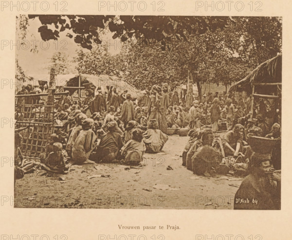 Women's pasar in Praja, Market in Praja, page, photomechanical print, maker: Christiaan Johan Neeb, c. 1892 - in or before 1897, Christiaan Johan Neeb, paper, collotype, photoprint, height 120 mm x width 167 mm, historical persons, market, Indonesia, A sepia-toned photograph showing a crowded outdoor scene with many people seated and standing on a dirt ground, many wearing loose garments and cloaks, some grouped around low bowls and containers, others sitting on the ground or on low stools, with simple thatched shelters and woven fences visible around the perimeter, large leafy trees overhead casting shade, scattered leaves on the ground, and a mix of adults and children occupying the space.