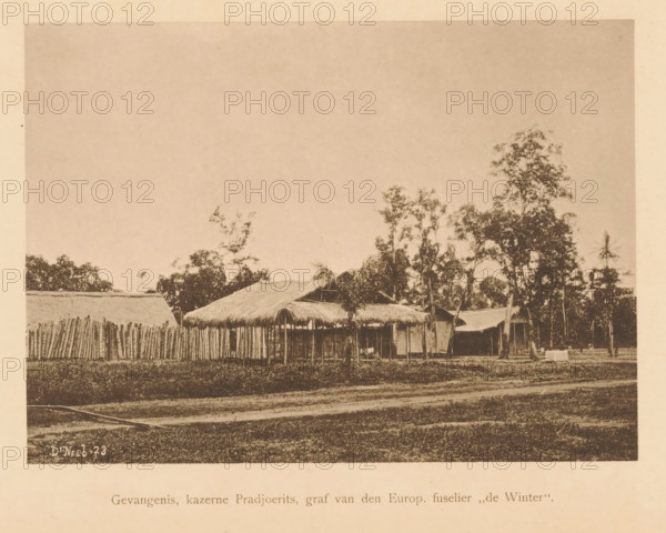Prison, Pradjoerits barracks, grave of the Europ. huselier 'de Winter', View of the Pradjoerits barracks, page, photomechanical print, maker: Christiaan Johan Neeb, c. 1892 - in or before 1897, paper, collotype, photoprint, height 121 mm x width 168 mm, barracks, Indonesia, A wide sepia-toned photograph showing a row of simple thatched-roof huts and a tall fence made of vertical wooden posts set behind a grassy foreground, with scattered trees of varying heights rising behind and between the structures and a broad expanse of pale sky above, the scene framed by a lighter border around the image.