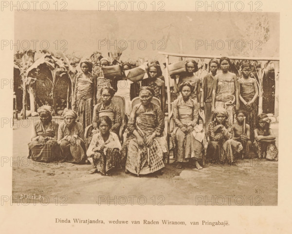 Dinda Wiratjandra, widow of Raden Wiranom, of Pringabaje, Portrait of unknown women and girls with Dinda Wiratjandra, page, photomechanical print, maker: Christiaan Johan Neeb, c. 1892 - in or before 1897, paper, collotype, photogravure, height 120 mm x width 165 mm, Person, historical persons, adult woman, adolescent, young woman, maiden, girl (child between toddler and youth), Indonesia, A sepia-toned photograph showing a group of people arranged in rows outdoors in front of tall leafy plants and a simple wooden structure, with several seated individuals wearing patterned cloth wrapped around their bodies and headscarves, and others standing behind them carrying woven baskets and bundles on their heads or shoulders; the seated row includes older women with folded hands and a few younger individuals seated on the ground, while the standing row shows women in wrapped garments and a couple of children, all facing the camera with neutral expressions and the ground in front of them visibl...