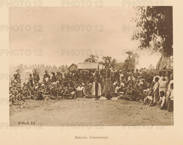 Balian Dancing Girls, Group of unknown persons watching two dancers, page, photomechanical print, maker: Christiaan Johan Neeb, c. 1892 - in or before 1897, paper, collotype, photoprint, height 120 mm x width 165 mm, adult man, dancing, anonymous historical persons depicted in a group, in a group-portrait, Indonesia, A wide sepia photograph shows a large crowd of people gathered outdoors with trees and simple thatched-roof huts in the background, many seated on the ground and others standing around a central open space where two women wearing patterned skirts and decorative tops stand facing forward, a few seated figures near them appear to hold drums or boxes, the crowd includes men, women, and children in traditional and simple clothing, and a large tree with hanging leaves is visible on the right edge of the scene.