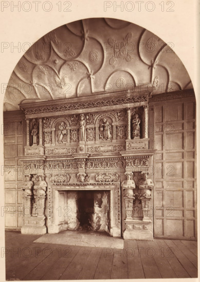 Fireplace at South Wraxall, Manor House Wilts., View of a mantelpiece at Manor House in South Wraxall, Wiltshire, page, photomechanical print, photographer: Foote, cliché maker: Woodbury Permanent Photographic Printing Company (possibly), c. 1871 - in or before 1876, paper, Woodburytype, height 117 mm x width 86 mm, open hearth, fire-place, A tall ornate stone fireplace with carved figures and decorative panels fills the lower portion of the image, flanked by carved pilasters topped with sculpted human busts and standing figures, and a large open firebox showing soot and darkened interior; above the mantel is an elaborately carved frieze with small sculpted figures set in arched niches and ornamental relief work, and the upper part of the image shows a curved ceiling with a repeating scalloped pattern and raised rosette and leaf motifs, while paneled wooden walling surrounds the fireplace and a wooden plank floor occupies the foreground.
