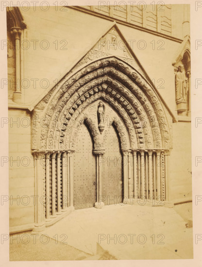 Portal of Lichfield Cathedral, photograph, page, photographer: John Benjamin Stone, Lichfield, c. 1865 - in or before 1870, Staffordshire, paper, albumen print, height 177 mm x width 128 mm, church (exterior), parts of church exterior and annexes: portal, parts of church exterior and annexes: tympanum, Lichfield Cathedral, A sepia-toned photograph of a carved stone doorway featuring a pointed arch with multiple concentric archivolts decorated with carved foliage and small sculpted heads, a central sculpted figure standing above double wooden doors studded with decorative rivets, slender clustered columns on either side with decorated capitals, flat stone walls surrounding the entrance, and a sculpted standing figure in a recessed niche to the right of the doorway.