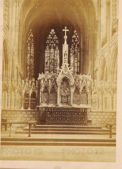 Interior of Lichfield Cathedral, photograph, page, photographer: John Benjamin Stone, Lichfield Cathedral, c. 1865 - in or before 1870, Staffordshire, Lichfield, paper, albumen print, height 180 mm x width 128 mm, interior of church, rood-screen, choir-screen, iconostasis, A sepia-toned interior view of a large gothic-style chapel showing an ornate altar area centered beneath tall pointed arches and stained glass windows. The altar features a carved reredos with pinnacles, arches, statues, and a prominent cross at its peak, and a decorative frontal with repeating motifs. Flanking the central structure are rows of slender columns and pointed arcades with tracery and carved figures. Behind the altar, tall lancet stained glass windows with patterned panes rise into the vaulted ceiling. In front of the altar there are stepped stone platforms and metal railings with twisted supports. The overall scene shows stone walls, detailed carvings, and high vaulted ribs forming the ceiling.