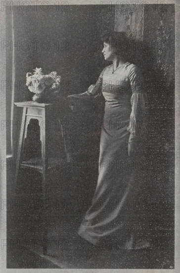 Portrait of an unknown woman, page, photomechanical print, inscription: 'Photographische Mitteilungen XL', monogram: 'BX', photographer: Fritz Loescher, cliché maker: unknown, c. 1898 - in or before 1903, paper, autotypie, height 190 mm x width 125 mm, historical persons - BB - woman, A grainy black-and-white photograph of a woman standing beside a narrow wooden table that holds a vase of flowers, the woman facing toward a window with her profile visible and her hand resting on the table, wearing a long, fitted dress with a decorative band across the chest and long fringed sleeves, her hair loose around her shoulders, patterned drapery or wallpaper visible behind her and soft light coming from the left illuminating the flowers and the side of her face.