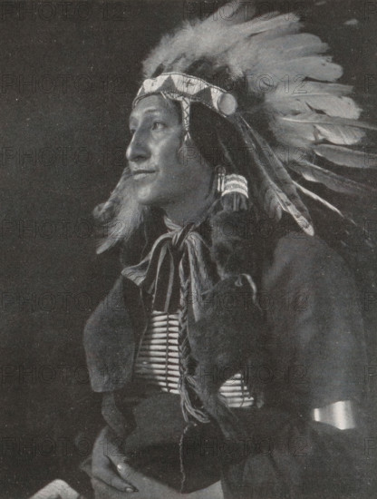 Portrait of an unknown man, The Black Fox, page, photomechanical print, monogram: 'MR&C', photographer: Gertrude Käsebier, New York (city), cliché maker: Meisenbach, Riffarth & Cie., c. 1898 - in or before 1903, North America, paper, autotypie, height 166 mm x width 129 mm, historical persons, A black and white portrait of a person wearing a feathered headdress with a decorative headband and long feathers extending backward, the person shown in three-quarter profile looking to the side, with long hair and braided elements, a beaded or bone chest ornament visible beneath layered clothing, fur or textured fabric draped over the shoulders, hands resting together in the lower frame, and a dark background surrounding the figure.
