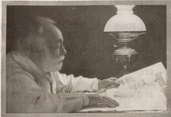 Bei der lampe, Portrait of an unknown man, page, photomechanical print, monogram: 'BX', inscription: 'Copenhagen', inscription: 'Photographische Mitteilungen XL', photographer: Niels Adam Fischer, cliché maker: unknown, c. 1898 - in or before 1903, paper, autotypie, height 125 mm x width 181 mm, historical persons, A grayscale image of an elderly man in profile leaning forward with a beard and glasses, wearing a loose garment and resting his forearms on a surface while holding a large folded paper, and a decorative lamp with an ornate base and a rounded glass shade positioned beside him.