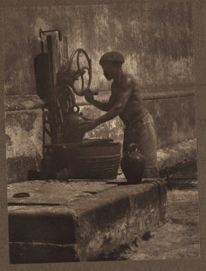 Water carrier in Cuba, A Water Carrier - Cuba, page, photomechanical print, photographer: Horace A. Latimer, cliché maker: anonymous, 1901, paper, photoaquatint, height 198 mm x width 147 mm, Print opposite title page, behind semi-transparent cover., adult man, pump, A sepia-toned photograph of a shirtless man wearing a cap and a cloth around his waist, standing beside a stone ledge and operating a hand crank attached to a vertical mechanical pump mounted on a wall; the man’s profile and muscular arms are visible as he leans toward a large basin beneath the pump, with a rounded ceramic jug placed on the ledge near his legs and weathered textured walls and stone surfaces surrounding the scene.