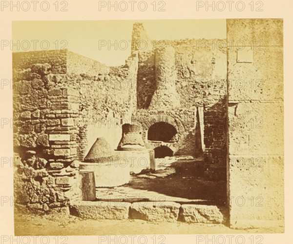 View of the ruins of the mill and ovens at Pompeii, photograph, page, Pompeii, c. 1862 - in or before 1867, photographer, paper, pasteboard, albumen print, height 139 mm x width 174 mm, Photo on third unnumbered page after page 38., ruin of a building, architecture, oven, kitchen - CC - in the open air, A sepia-toned photograph of a stone ruin featuring rough textured walls and stacked masonry, with a passageway leading toward an arched opening set into a brick-faced recess; in the foreground there are cylindrical and conical stone structures placed on square bases, a paved stone floor with worn blocks, and a tall upright stone slab casting a shadow; the right edge shows a smooth, vertically jointed stone surface contrasting with the rubble masonry, and the upper area reveals a crumbling wallline against a pale sky.