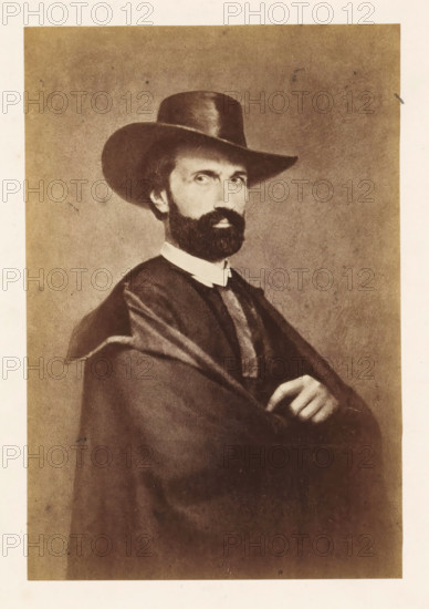 Portrait of Antoine-Joseph Wiertz, photograph, page, photographer: attributed to Edmond Fierlants, c. 1855 - in or before 1865, Attributed to, paper, albumen print, height 169 mm x width 114 mm, Photo opposite title page., Person, Antoine Joseph Wiertz, historical persons, A sepia-toned portrait of a man shown from the waist up wearing a wide-brimmed hat and a dark cloak draped over his shoulders, with a white collar visible at his neck; he has a full dark beard and mustache, short hair peeking beneath the hat, and a serious expression while his arms are crossed with one hand resting on the other.