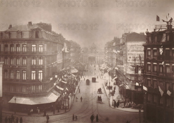 Lille, vue de la Rue de la Gare, View of the Rue Faidherbe with the train station in Lille, page, photomechanical print, inscription: 'Le Nord Contemporain, Revue Artistique & Archéologique', photographer: Lemercier, cliché maker: Mallart, c. 1878 - in or before 1882, paper, Woodburytype, height 151 mm x width 212 mm, Print on third unnumbered page after page 240., city-view in general, 'veduta', street, A wide street scene viewed from above shows rows of multi-story buildings with mansard roofs and dormer windows lining both sides, shop awnings and flags projecting over the sidewalks, scaffolding and rooftop structures on some buildings, and a crowd of people on the sidewalks and walking in the street. Horse-drawn carriages and a few early automobiles move along tracks embedded in the pavement, while lampposts stand along the roadway. The far end of the street is framed by a large archway or gateway between buildings, and the sky above is overcast.