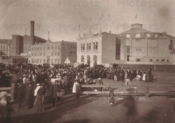 Roubaix, l'Hippodrome a la sortie des sociétés chorales, View of the Grand Place in Roubaix with visitors and a church choir, page, photomechanical print, inscription: 'Pendant les fêtes des 13, 14 et 15 Août', inscription: 'Le Nord Contemporain, Revue Pittoresque', photographer: Lemercier, cliché maker: Ferrand, c. 1878 - in or before 1882, Lemercier, Ferrand, photographer, cliché maker, paper, Woodburytype, height 151 mm x width 214 mm, Print on third unnumbered page after page 272., square, circus, choir, A wide sepia photograph shows a crowded outdoor scene in front of large brick buildings with varied architectural details; a grand façade with arched windows and decorative pinnacles stands near a round building with a domed roof and a flag on top, while adjacent structures include a tall tower and multi-story brick blocks. The foreground is filled with many people wearing period clothing, including hats, coats, long dresses, and aprons, some pushing or standing near small wheeled ...