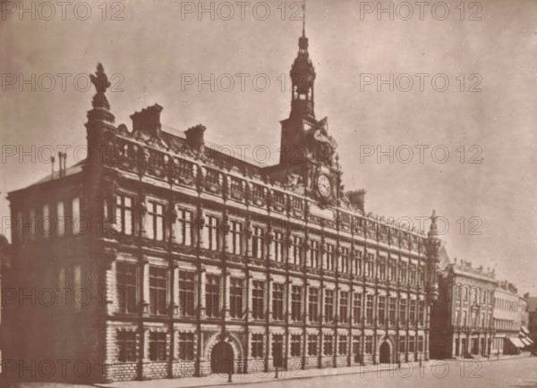 View of the Valenciennes town hall, L'Hotel-de-Ville de Valenciennes, page, photomechanical print, inscription: 'Le Nord Contemporain, Revue Artistique & Archéologique', photographer: Lemercier, Valenciennes, cliché maker: Lemercier, c. 1878 - in or before 1882, Lemercier, photographer, cliché maker, paper, Woodburytype, Woodburytypepie, height 142 mm x width 194 mm, Print on third unnumbered page after page 48., townhall, A sepia-toned photograph of a large ornate building seen from an angle showing its front and side façades, with multiple rows of tall rectangular windows set in decorative stonework and arched doorways at street level, a detailed cornice and sculptural roofline with statues and ornamental elements, and a central tower rising above the roof topped by a spire and housing a circular clock face; the street in front is mostly empty with a few indistinct figures near the building and adjacent lower buildings extending along the right side of the image.