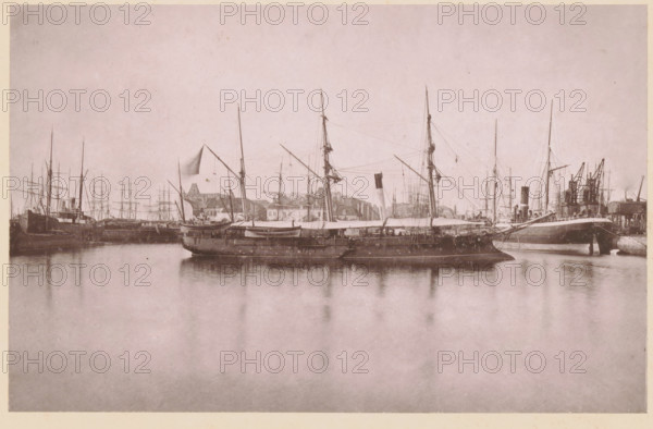 La Lionne, Canonnière de l'État, View of La Lionne near Calais, a gunboat of France, page, photomechanical print, inscription: '(en rade de Calais)', inscription: 'Le Nord Contemporain, Revue Pittoresque', photographer: NC, Calais, cliché maker: Luzzato, Dunkirk, c. 1877 - in or before 1882-12-15, NC, Luzzato, photographer, cliché maker, paper, collotype, photoprint, height 160 mm x width 251 mm, Print on third unnumbered page after page 368., sailing-ship, sailing-boat, firearms: cannon, A wide harbor scene showing several sailing vessels and steamships moored along a quay, with tall masts, rigging, and spars rising against a pale sky, a larger ship in the foreground with lifeboats and canvas covers on its deck, reflections of the ships visible on calm water, a line of warehouses and cranes along the shore behind the vessels, and an overall muted, monochrome tone.