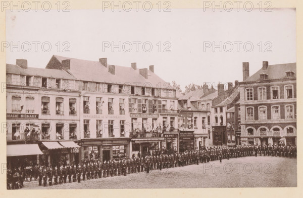 View of the Grand'Place in Maubeuge, France, Maubeuge, La Grand'Place, page, photomechanical print, inscription: 'Le Nord Contemporain, Revue Artistique & Archéologique', photographer: NC, cliché maker: Auguste Chapuis, c. 1877 - in or before 1882-12-15, paper, collotype, photoprint, height 162 mm x width 262 mm, Print on fifth unnumbered page after page 368., square, circus, A wide cobblestone street in front of a row of multi-story brick and stone buildings with many windows and balconies, several awnings over ground-level shopfronts, and people standing or sitting at windows and on balconies holding small flags; a large formation of uniformed soldiers wearing helmets and carrying rifles lines the street in orderly ranks stretching across the foreground while a single officer stands apart facing them; the buildings show varied rooflines with chimneys, dormer windows, and decorative facades, and a crowd of onlookers is gathered near the shop entrances.