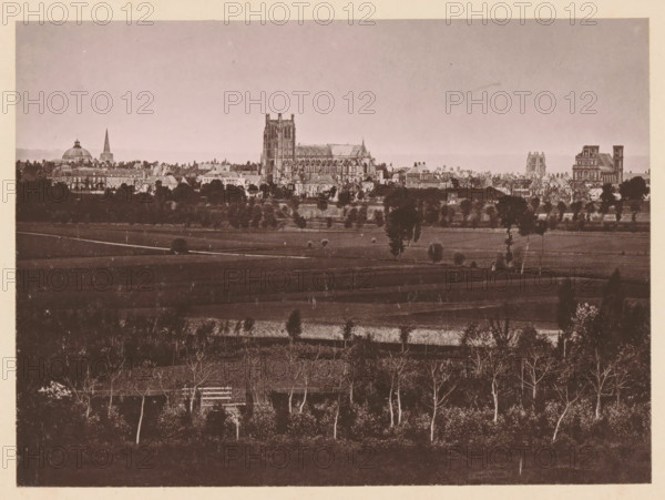 Vue Générale de Saint-Omer, View of Saint-Omer, page, photomechanical print, inscription: 'Le Nord Contemporain, Revue Historique & Pittoresque', photographer: NC, Saint-Omer, cliché maker: C. Becquereau, c. 1878 - in or before 1883-02-05, paper, collotype, photoprint, height 175 mm x width 238 mm, Print on fifth unnumbered page after page 32., prospect of city, town panorama, silhouette of city, A wide sepia-toned cityscape seen from a distance with a foreground of darker fields and rows of leafless trees, a midground of lighter cultivated land with scattered trees and a few small structures, and a background skyline dominated by large historic buildings including a cathedral with tall towers and intricate stonework, a domed building, a slender spire, and several multi-story stone structures, all beneath a pale sky.