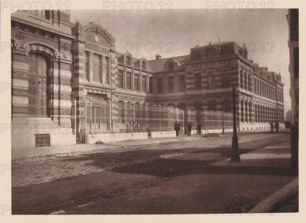 Lille, La Nouvelle Faculté de Médicine, View of Place de la République in Lille during a market, page, photomechanical print, inscription: 'Le Nord Contemporain, Revue Artistique & Archéologique', inscription: 'Elevée par MC Batteur, Architecte', inscription: 'Journal le Nord Contemporain, No 18, année 1882.', photographer: Lemercier, cliché maker: Ferrand, c. 1877 - in or before 1882-09-30, paper, Woodburytype, height 152 mm x width 212 mm, Print on fifth unnumbered page after page 288., market, university (building, education, tuition), A sepia-toned photograph of a large ornate brick and stone building with tall arched windows, decorative cornices, and a clock set in an elaborately carved pediment near the roofline, a metal fence with ornate vertical bars runs along the sidewalk in front of the building, several people stand or walk near the fence and along the cobblestone street, a lamppost stands near the curb casting a shadow on the pavement, the foreground shows a rough dirt and...
