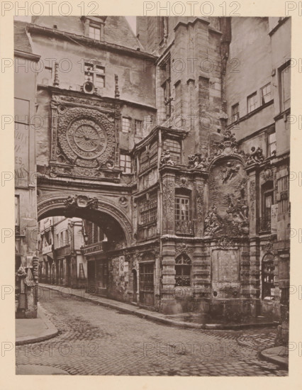 Rouen, La Grosse Horloge, View of the Gros-Horloge in Rouen, page, photomechanical print, inscription: 'Le Nord Contemporain, Revue Artistique & Archéologique', inscription: 'Journal le Nord Contemporain, No 27, année 1883.', photographer: NC, c. 1878 - in or before 1883-02-20, paper, collotype, photoprint, height 221 mm x width 168 mm, Print on third unnumbered page after page 48., tower, clock tower, monumental adornment, Gros-Horloge, A narrow cobblestone street lined with tall, weathered buildings leads under a large ornate stone archway attached to a facade featuring an elaborate circular clock surrounded by carved floral and scrollwork motifs. Above and beside the arch are multi-paned windows and sculpted decorative elements including relief panels with figures and cherubs, carved pilasters, and balustraded window boxes. The stone surfaces show aged textures and darkened areas, and a small inset doorway and arched window occupy the ground level beneath the relief. A portion of th...