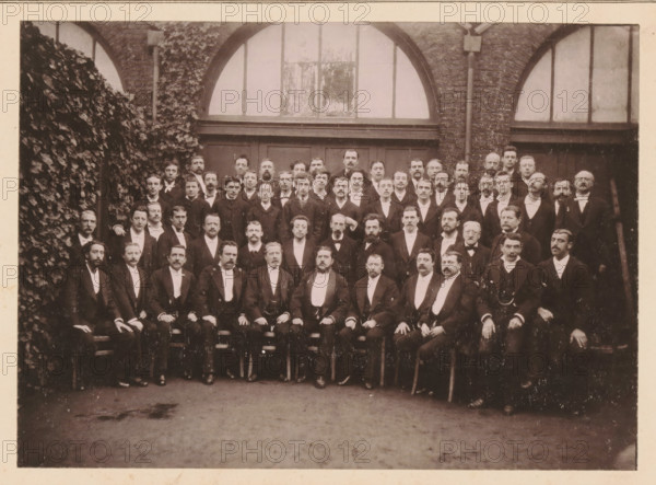 L'Orchestre, de la Société des Concerts Popculaires de Lille, Group portrait of the orchestra of the Société des Concerts Popculaires in Lille, page, photomechanical print, inscription: 'Le Nord Contemporain, Revue Artistique', photographer: NC, Lille (possibly), cliché maker: Ferrand, c. 1878 - in or before 1883-03-20, NC, Ferrand, photographer, cliché maker, Lille, paper, collotype, photoprint, height 167 mm x width 231 mm, Print on fifth unnumbered page after page 80., large group of musicians, orchestra, anonymous historical persons depicted in a group, in a group-portrait, A large group of men arranged in several rows, most seated in the front and standing behind, all wearing dark formal suits with light shirts and ties, many with mustaches and beards, their hands resting on laps or knees; they are positioned in front of a brick building featuring large arched windows and a wooden doorway, with a dense wall of climbing ivy visible along the left side and a vertical drainpipe near ...