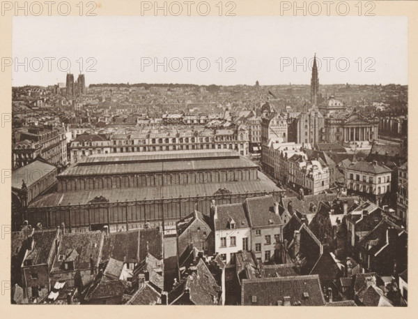 View of Brussels, Bruxelles, Vue Générale, page, photomechanical print, inscription: 'Journal le Nord Contemporain, No 34, année 1883.', inscription: 'Le Nord Contemporain, Revue Artistique & Archéologique', photographer: NC, Brussels, c. 1878 - in or before 1883-06-05, paper, collotype, photoprint, height 160 mm x width 220 mm, Print on third unnumbered page after page 160., prospect of city, town panorama, silhouette of city, A wide aerial view of an urban area showing many tightly packed buildings with varied rooflines and tiled roofs in the foreground, a large long building with a metal or glass roof and a colonnaded facade occupying the midground, rows of multi-story masonry buildings with ornate façades and dormer windows behind it, and a distant cityscape with taller church spires and towers rising above the lower rooftops under a pale sky.
