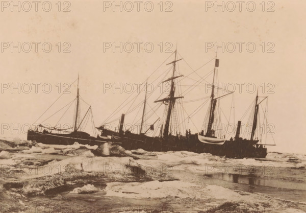 The Dymphna and the Varna shortly after freezing. September 23 1882., View of the Dymphna and the Varna in the Kara Sea, page, photomechanical print, inscription: 'De Nederlandsche Polar-Expeditie 1882-1883.', photographer: H. Ekama, Karskoye More, cliché maker: E. Diener, Glauchau, c. 1881 - in or before 1886, Saxony, paper, collotype, photoprint, height 140 mm x width 206 mm, ships (in general), landscapes in polar regions, A sepia-toned scene showing several docked sailing ships viewed from the side, with tall masts, rigging, and spars silhouetted against a pale sky, small lifeboats hanging from davits on one vessel, decks with indistinct figures and equipment, a rocky foreground with ice floes and textured water, and decorative border details along the top edge of the image.