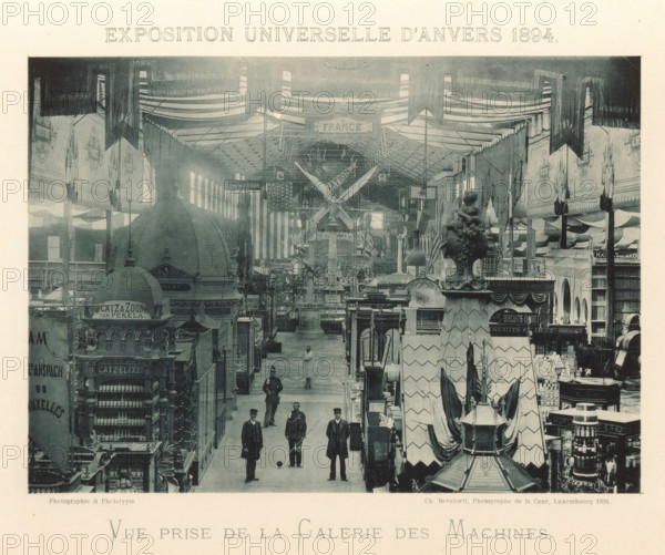 View of the machine gallery of the Antwerp World Exhibition in 1894, Vue prize de la Galerie des Machines, Exposition Universelle d'Anvers 1894, page, photomechanical print, photographer: Charles Bernhoeft, Antwerp, cliché maker: Charles Bernhoeft, Luxembourg (Belgian province), 1894, Charles Bernhoeft, photographer, cliché maker, paper, collotype, photoprint, height 154 mm x width 216 mm, Print on third unnumbered page after page 26., market-hall, world fair, world exhibition, machines, parts of machines, tools and appliances, World Exhibition 1894 (Antwerp), Period, 1894-05-05 - 1894-11-05, A large indoor exhibition hall filled with rows of ornate booths and decorative kiosks beneath a high ceiling draped with striped fabric and hanging banners, with a central aisle leading into the distance. Several uniformed men stand and walk along the aisle, and a person in a white coat is visible further back. Sculptural architectural elements and ornamental finials rise above some booths, and a...