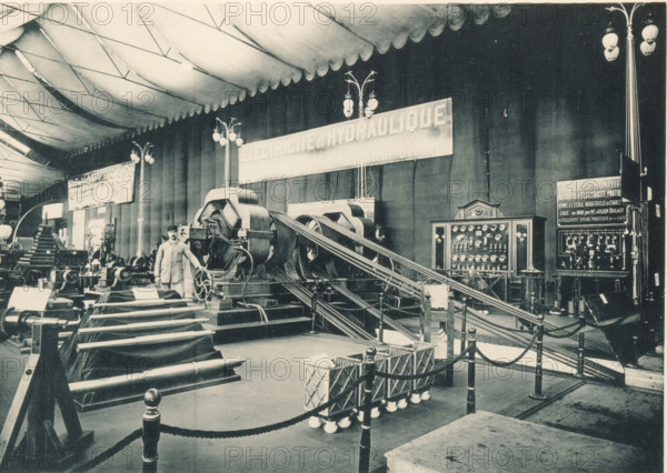 View of a setup of the Société Anonyme Électricté et Hydraulique from Charleroi at the Antwerp World Exhibition in 1894, Électricité et Hydraulique Charleroi, Exposition Universelle d'Anvers 1894, page, photomechanical print, photographer: Charles Bernhoeft, Antwerp, cliché maker: Charles Bernhoeft, Luxembourg (Belgian province), 1894, Charles Bernhoeft, photographer, cliché maker, paper, collotype, photoprint, height 153 mm x width 216 mm, Print on third unnumbered page after page 30., world fair, world exhibition, fluid mechanics, hydraulics, electrical phenomena, World Exhibition 1894 (Antwerp), Period, 1894-05-05 - 1894-11-05, A large indoor exhibition space with a high scalloped fabric ceiling and tall paneled walls, filled with industrial machinery and display equipment. In the foreground there are rope barriers attached to posts surrounding a collection of cylindrical components and framed coils mounted on low stands. A set of heavy belts or cables run from large mechanical drum...