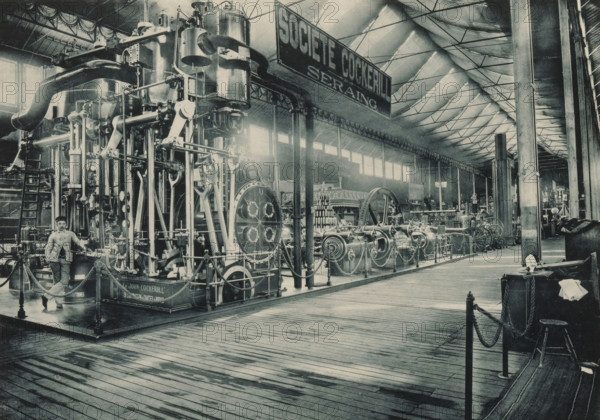 Société John Cockerill Seraing, View of a setup of Société Anonyme John Cockerill from Seraing at the Antwerp World Exhibition in 1894, Exposition Universelle d'Anvers 1894, page, photomechanical print, photographer: Charles Bernhoeft, Antwerp, cliché maker: Charles Bernhoeft, Luxembourg (Belgian province), 1894, Charles Bernhoeft, photographer, cliché maker, paper, collotype, photoprint, height 153 mm x width 216 mm, industries, metal, world fair, world exhibition, machines, parts of machines, tools and appliances, World Exhibition 1894 (Antwerp), Period, 1894-05-05 - 1894-11-05, A large indoor exhibition space with a high pitched ceiling supported by metal trusses and tall columns, a long wooden plank floor in the foreground, and an extensive display of heavy industrial machinery occupying the left and center areas. The machinery includes tall vertical cylinders, large flywheels, connecting rods, pipes, valves, chains, and ornate metal frameworks mounted on raised platforms with rope...