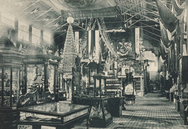 Coup d'oeil sur la section Allemande, Galerie Principale, View of the German part of the main exhibition of the Antwerp World Exhibition in 1894, Exposition Universelle d'Anvers 1894, page, photomechanical print, photographer: Charles Bernhoeft, Antwerp, Luxembourg (Belgian province), 1894, paper, collotype, photoprint, height 152 mm x width 222 mm, counter, shop, world fair, world exhibition, World Exhibition 1894 (Antwerp), Period, 1894-05-05 - 1894-11-05, A large indoor exhibition space filled with ornate wooden display cases, shelves, and decorative booths arranged on a patterned wooden floor. Tall vertical banners hang from the ceiling alongside draped fabric and structural beams, and a high roof with visible rafters and large windows lets light into the hall. Central displays include a tall conical tower made of stacked items topped with a spherical object, rows of bottles and containers on shelves, sculptural figures and carved pedestals, and glass-topped cabinets showing items ...