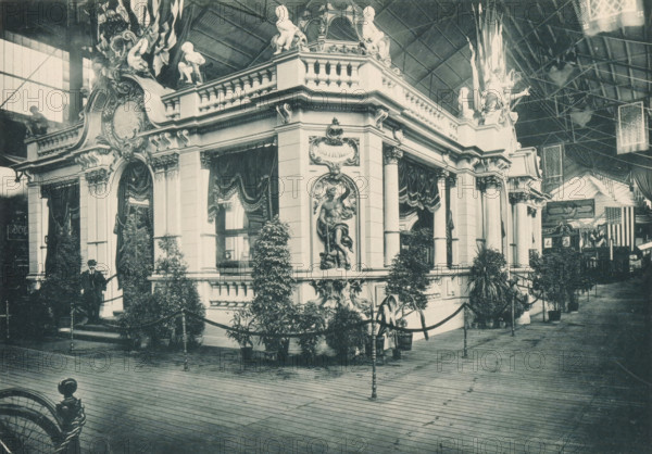 View of the Norddeutscher Lloyd pavilion at the Antwerp World Exhibition in 1894, Pavillon du Norddeutscher Lloyd, Exposition Universelle d'Anvers 1894, page, photomechanical print, photographer: Charles Bernhoeft, Antwerp, cliché maker: Charles Bernhoeft, Luxembourg (Belgian province), 1894, Charles Bernhoeft, photographer, cliché maker, paper, collotype, photoprint, height 153 mm x width 217 mm, palace, world fair, world exhibition, World Exhibition 1894 (Antwerp), Period, 1894-05-05 - 1894-11-05, A large ornate pavilion with classical architectural details occupies the center of the image, featuring columns, balustrades, decorative reliefs, and sculpted figures on its roofline; draped curtains hang over several openings and plants in pots line the exterior, with a rope barrier and stanchions bordering the structure; a man in a dark suit and hat stands near one entrance on a small set of steps; the scene is set inside a spacious exhibition hall with a high arched roof and visible met...