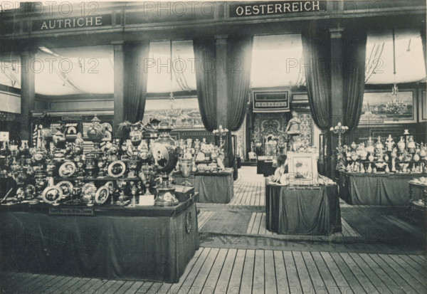 View of the Austrian part of the Antwerp World Exhibition in 1894, Section Autrichienne, Exposition Universelle d'Anvers 1894, page, photomechanical print, photographer: Charles Bernhoeft, Antwerp, cliché maker: Charles Bernhoeft, Luxembourg (Belgian province), 1894, Charles Bernhoeft, photographer, cliché maker, paper, collotype, photoprint, height 153 mm x width 222 mm, Print on third unnumbered page after page 42., counter, shop, container of ceramics: jar, jug, pot, vase, dish, plate, saucer, world fair, world exhibition, World Exhibition 1894 (Antwerp), Period, 1894-05-05 - 1894-11-05, A wide interior scene shows multiple display tables covered with dark cloths holding numerous ornate vases, plates, urns, figurines, and decorative objects arranged closely together, with a large decorative urn placed prominently in the foreground. Tall columns rise toward the ceiling between heavy draped curtains that frame an open central passage leading to another room where a statue stands. Chan...